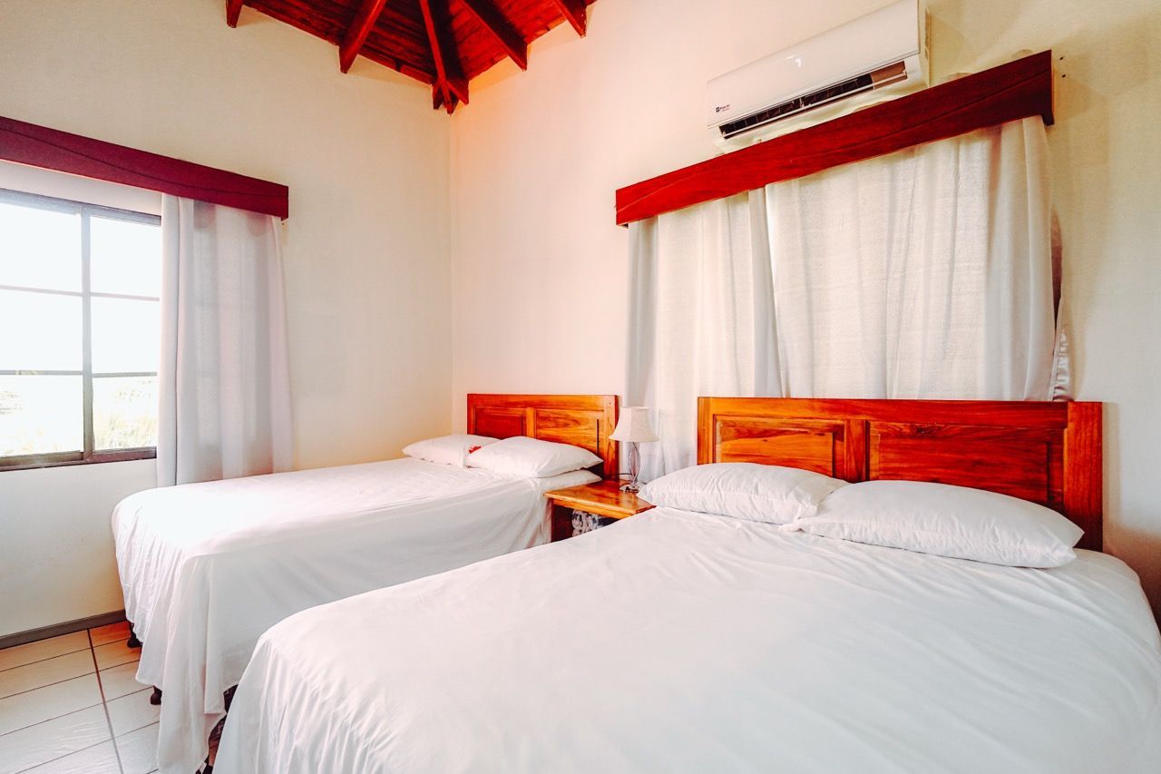 Dormitorio con dos camas, sábanas blancas y cabeceros de madera, con ventana y equipo de aire acondicionado.