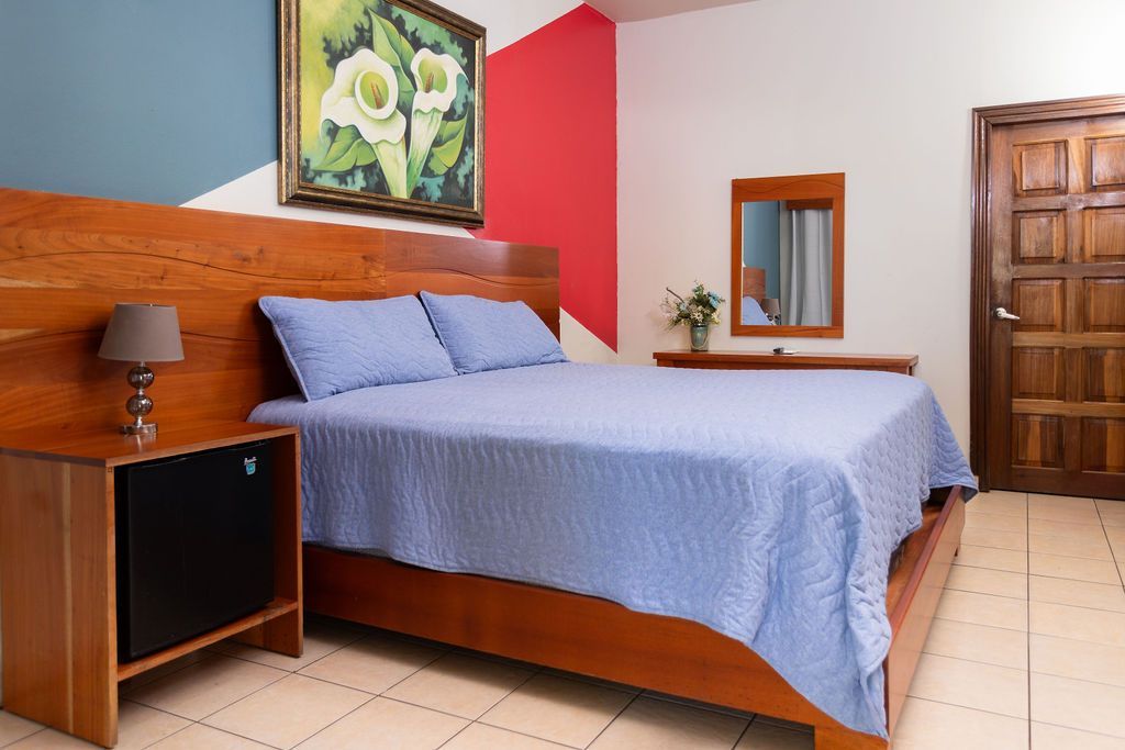 Dormitorio con cama, mesita de noche, cuadro, espejo y puerta de madera. Paredes azules, rojas y blancas.