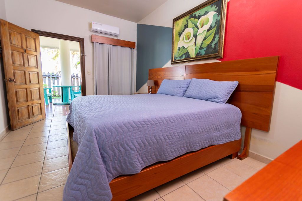 Dormitorio con cama, obras de arte y puerta abierta al balcón. Ropa de cama azul claro, cabecero y ribete de madera.