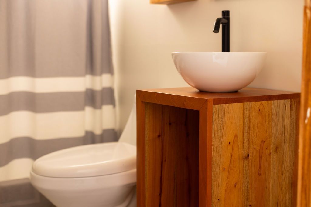 Baño con lavabo blanco sobre mueble de madera, grifo negro, inodoro y cortina de ducha a rayas.