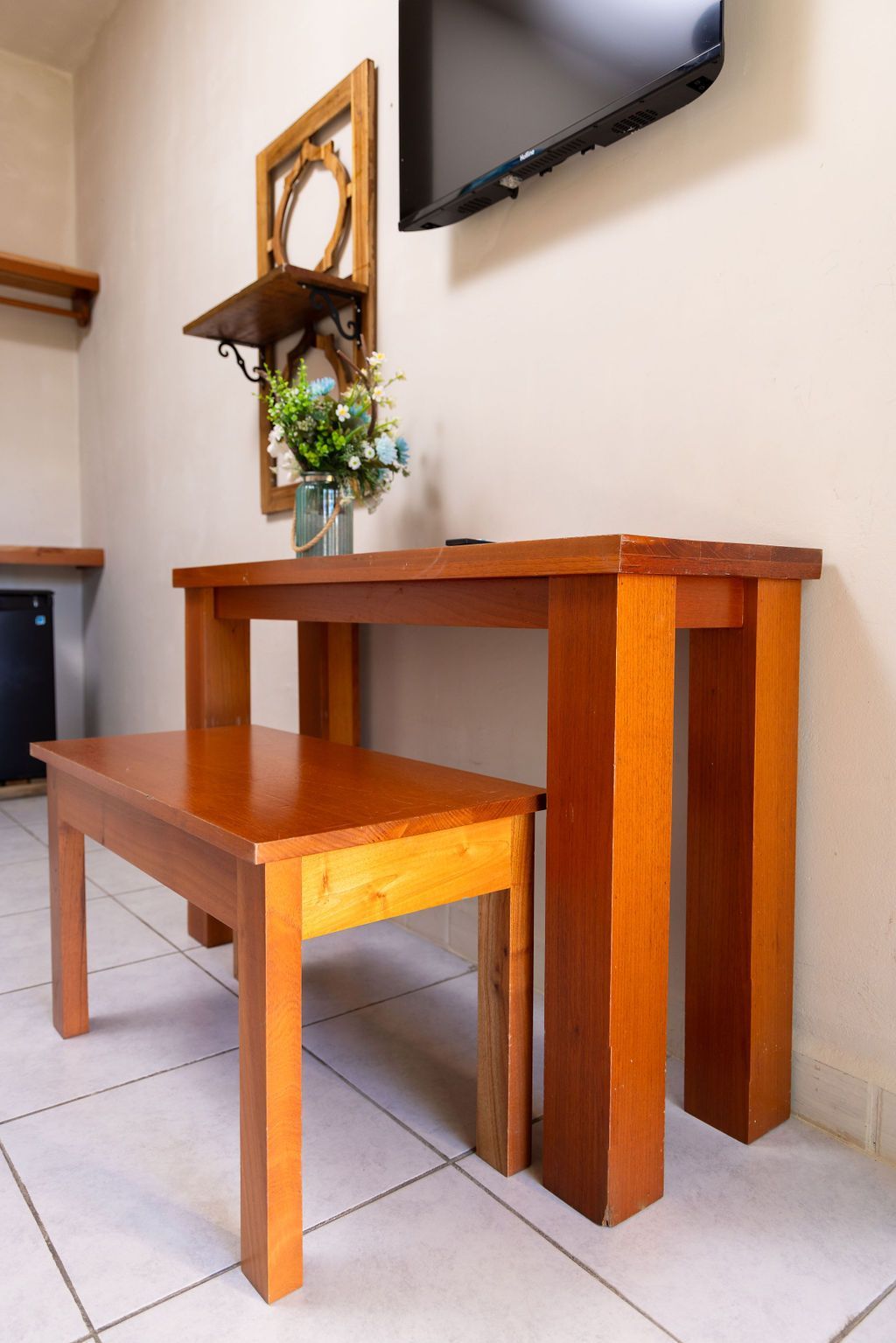 Mesa y banco de madera con flores, debajo de un televisor colgado, en una habitación con un estante