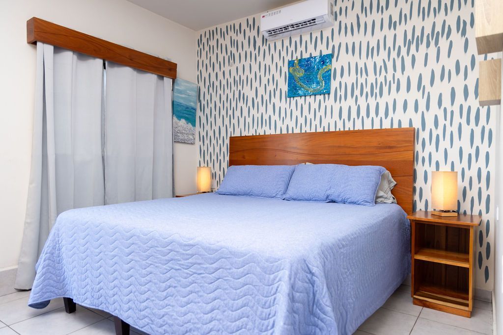 Dormitorio con cama azul, papel pintado estampado, cabecero de madera, mesita de noche con lámparas y cortinas.
