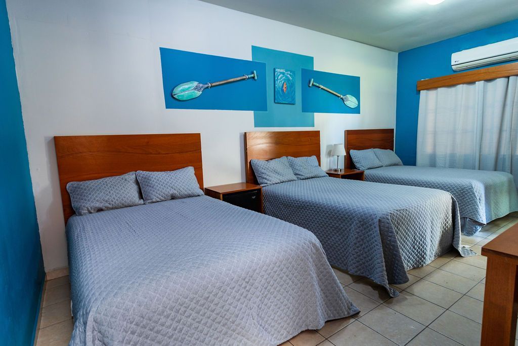 Tres camas en una habitación de hotel con paredes azules y blancas, obras de arte y una mesita de noche de madera.