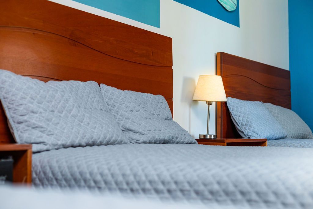 Dos camas con ropa de cama gris, cabeceros de madera y una lámpara en una habitación con detalles azules.