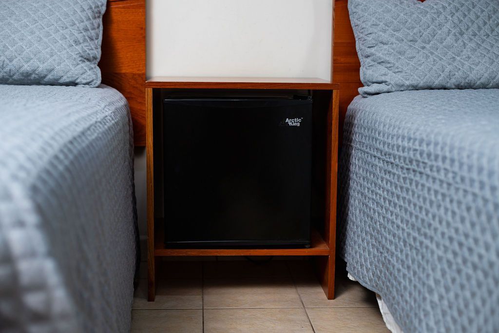 Mini-nevera en estante de madera entre dos camas con colchas y almohadas azules.