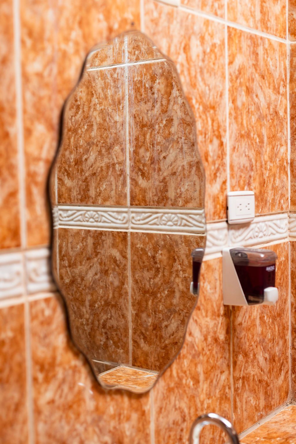 Espejo en pared de azulejos, dispensador de jabón a la derecha. Azulejos marrones y blancos.