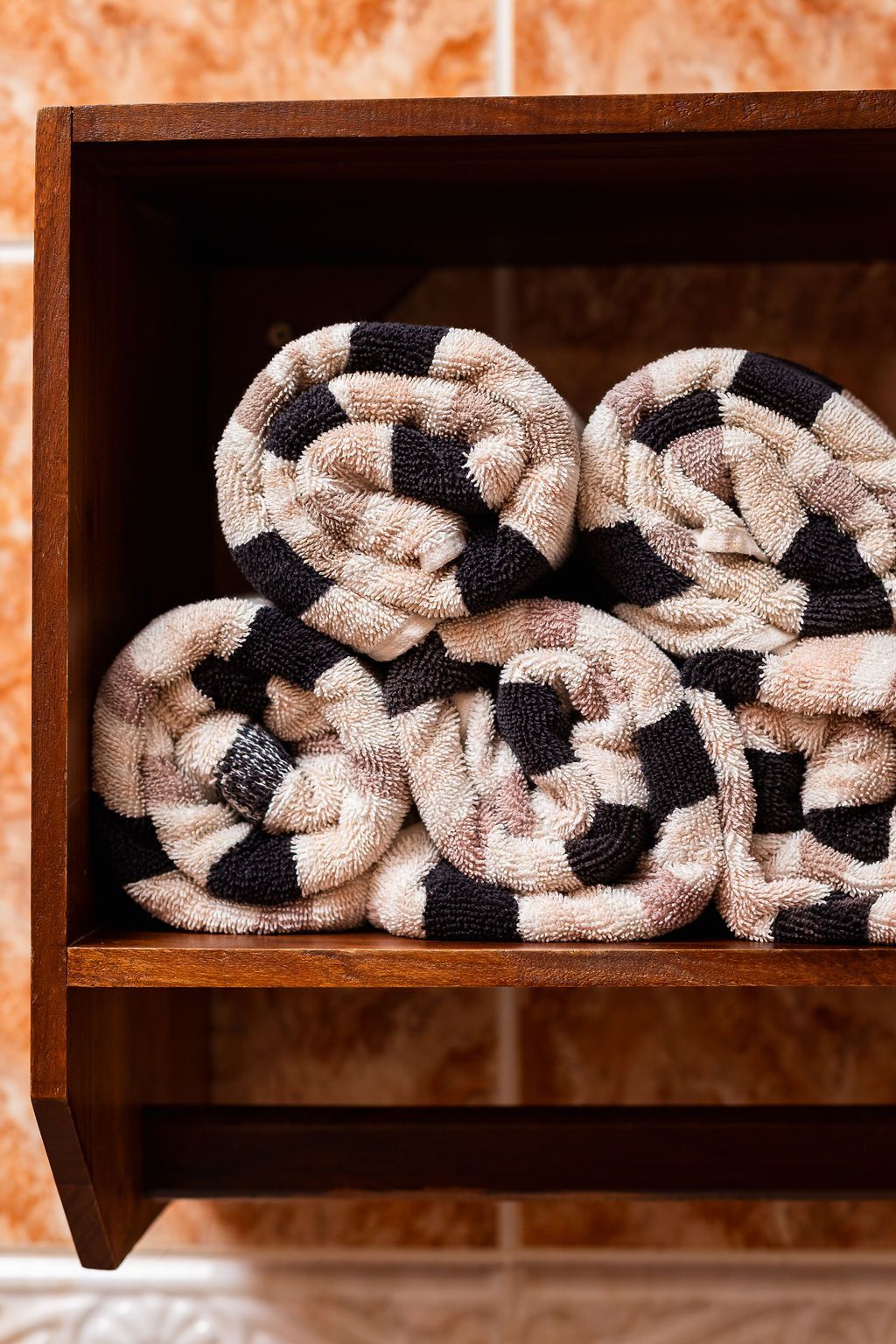 Estante de madera marrón que contiene seis toallas estampadas enrolladas sobre un fondo de azulejos.