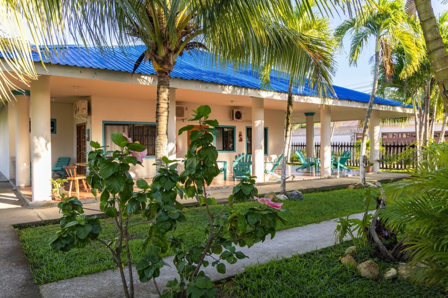 Un edificio blanco con techo azul y porche, bordeado de plantas verdes y palmeras.
