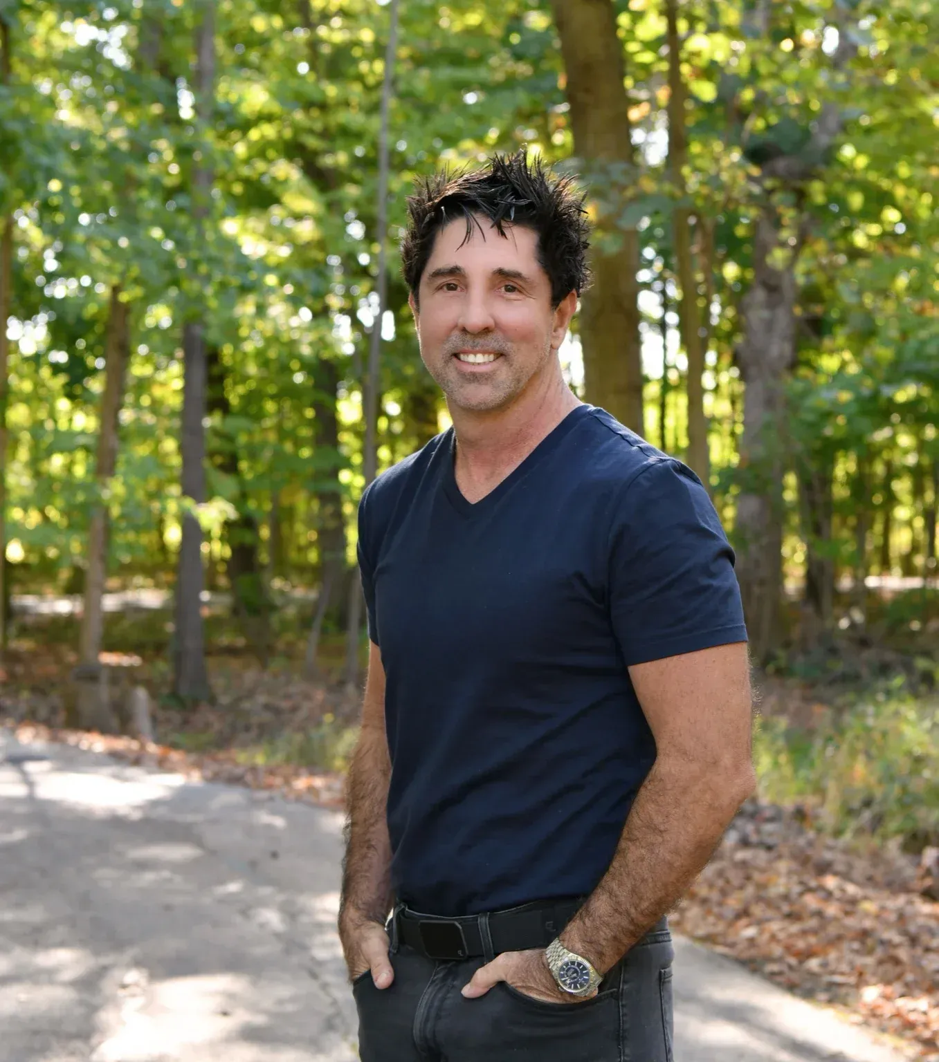 Man in a black suit with a gold tie, smiling with hands clasped, against a dark backdrop.