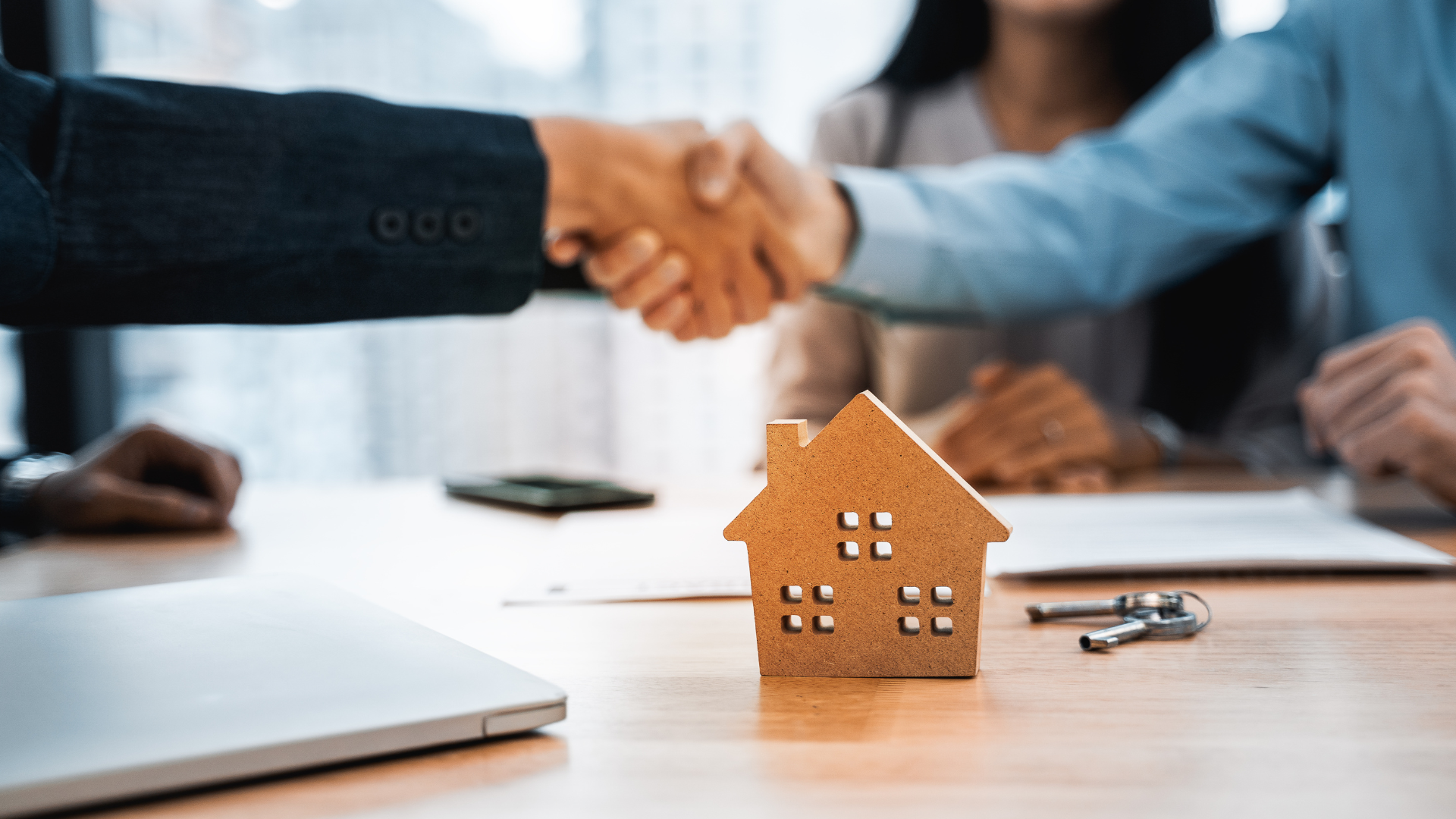 Two people shake hands over a table with a wooden house model and keys, signaling a successful real estate transaction.