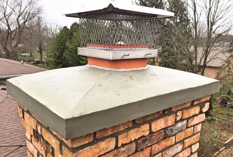 Brick chimney with concrete cap and metal mesh chimney cap in an outdoor setting.