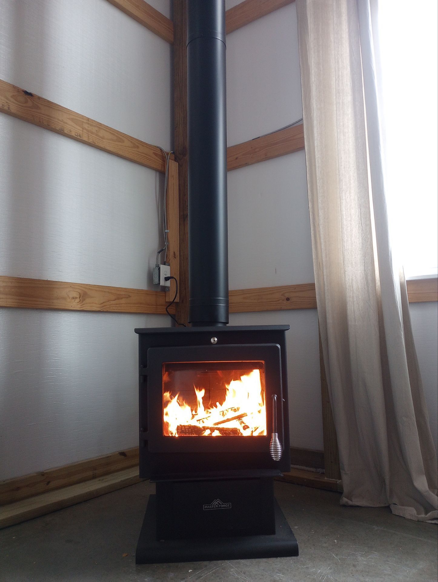 Wood-burning stove with flames in a corner of a room, black chimney, neutral walls and curtain.
