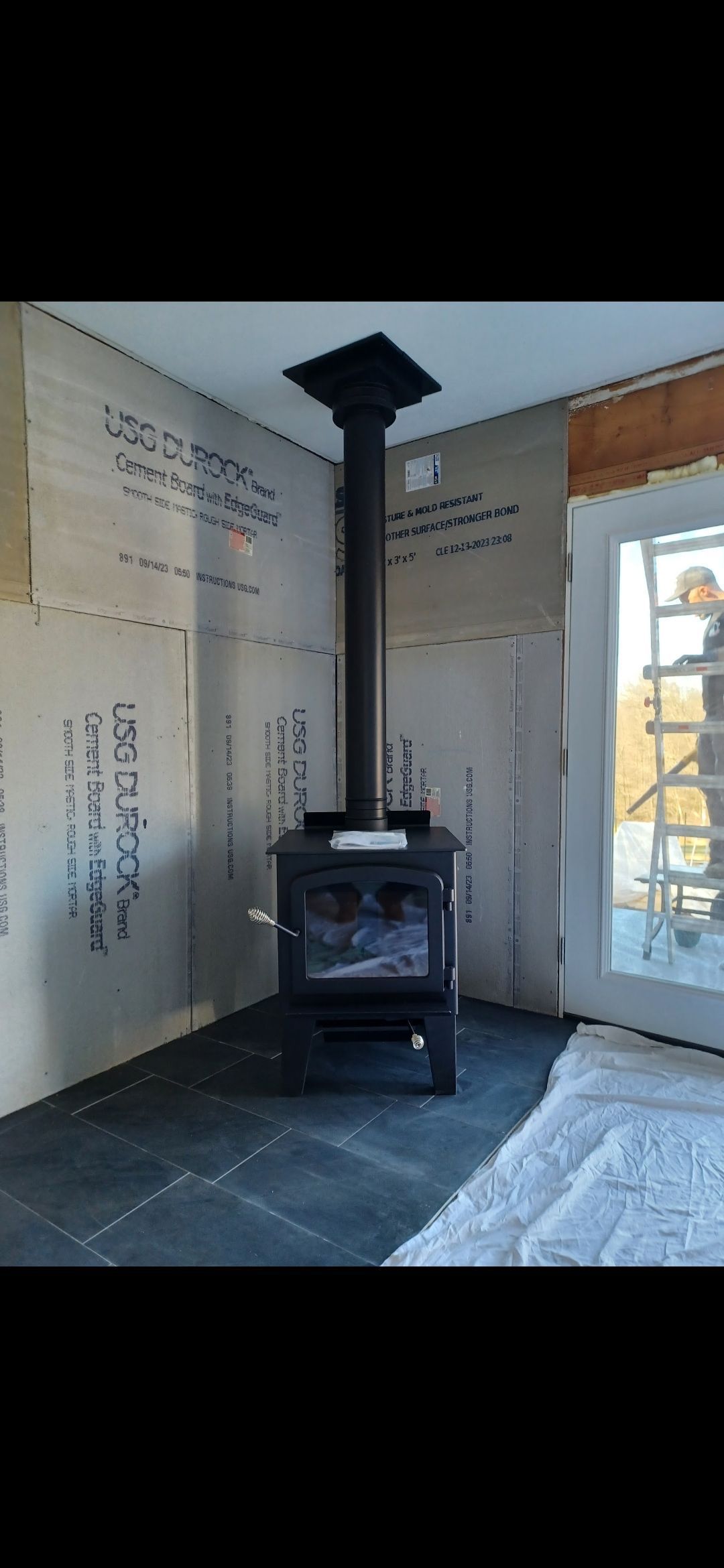 Black wood-burning stove with black chimney pipe, installed in a room with light-colored walls and dark floor tiles.