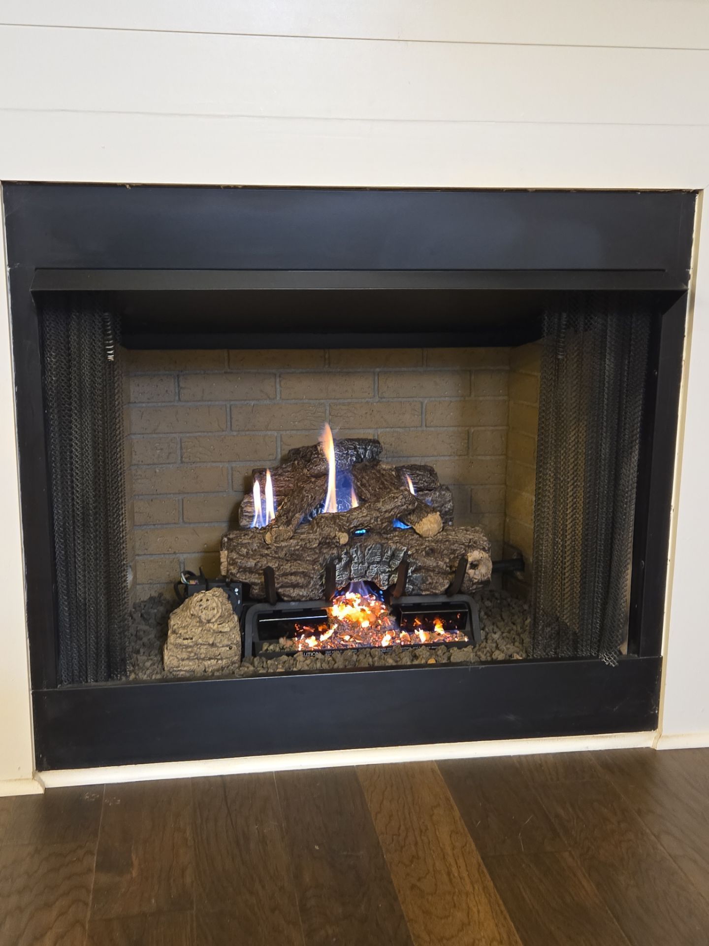Gas fireplace with burning logs and flames, set in a black frame, against a brick background.