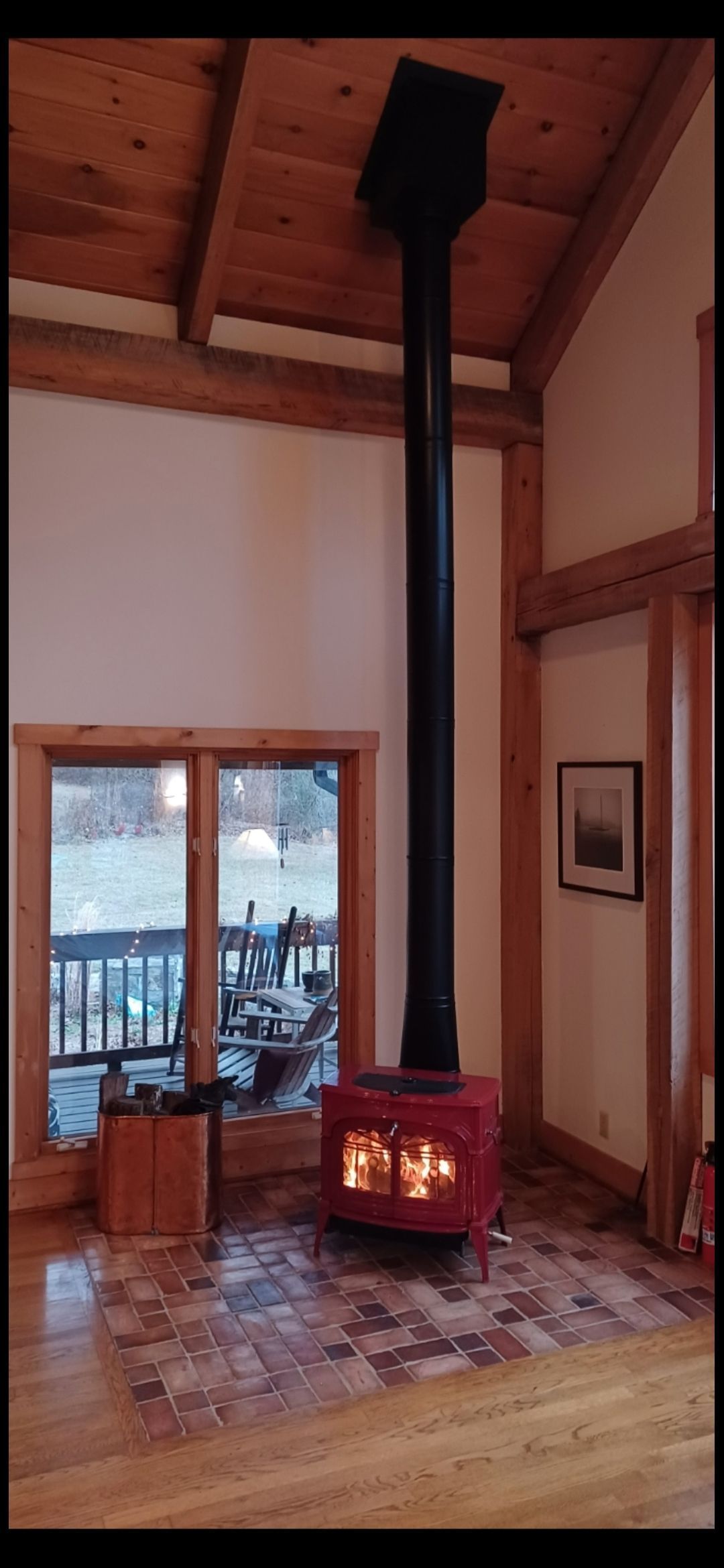 A red wood-burning stove with a black chimney in a cozy room with wooden beams and a window overlooking a deck.