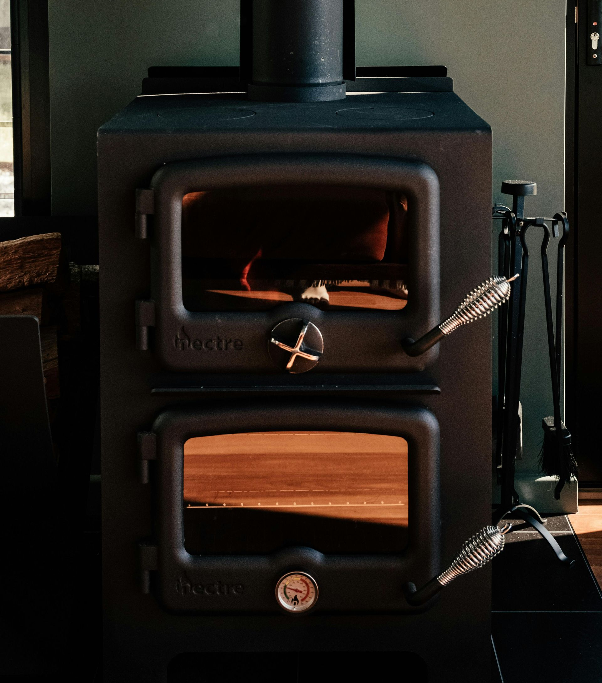 Black wood-burning stove with two glass doors, burning wood. Handles and tools are on the side.