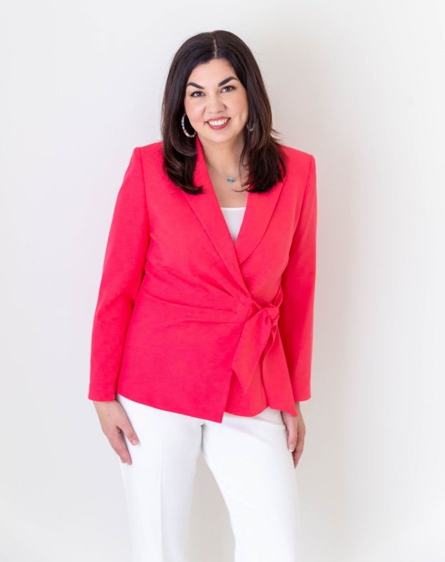 A woman in a red jacket and white pants is standing in front of a white wall.