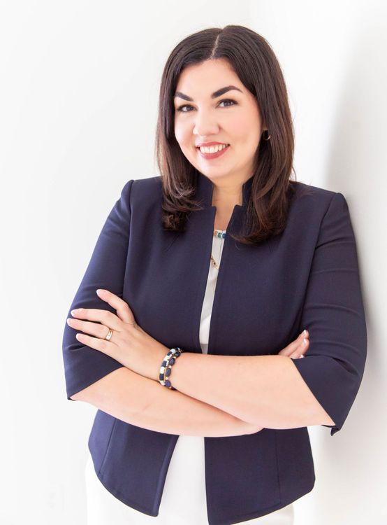 A woman in a blue jacket is standing with her arms crossed and smiling.