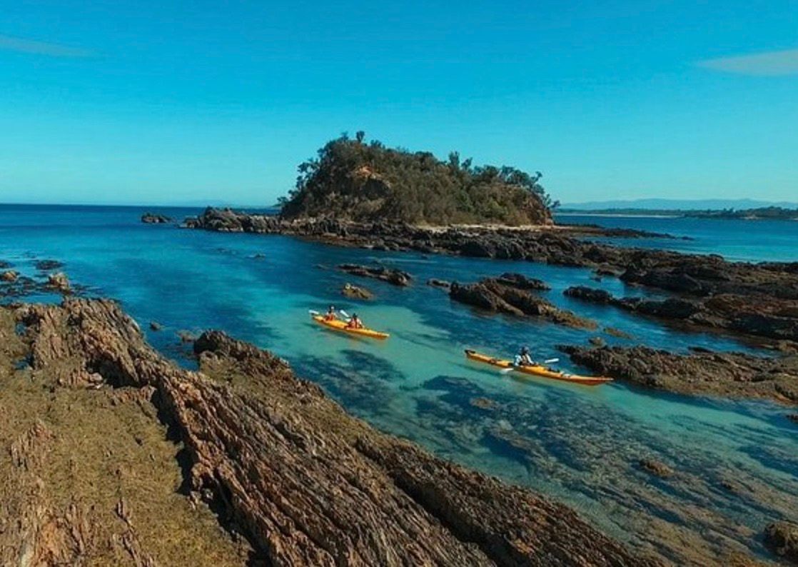 Two yellow kayaks in a rocky cove at Batemans Bay, South Coast, New South Wales  This image shows two yellow kayaks in a rocky cove at Batemans Bay, a popular tourist destination on the South Coast of New South Wales, Australia. The kayaks are resting on the shore of the cove, which is backdropped by a rocky island with lush vegetation. The water in the cove is calm and clear, and the sky is blue.