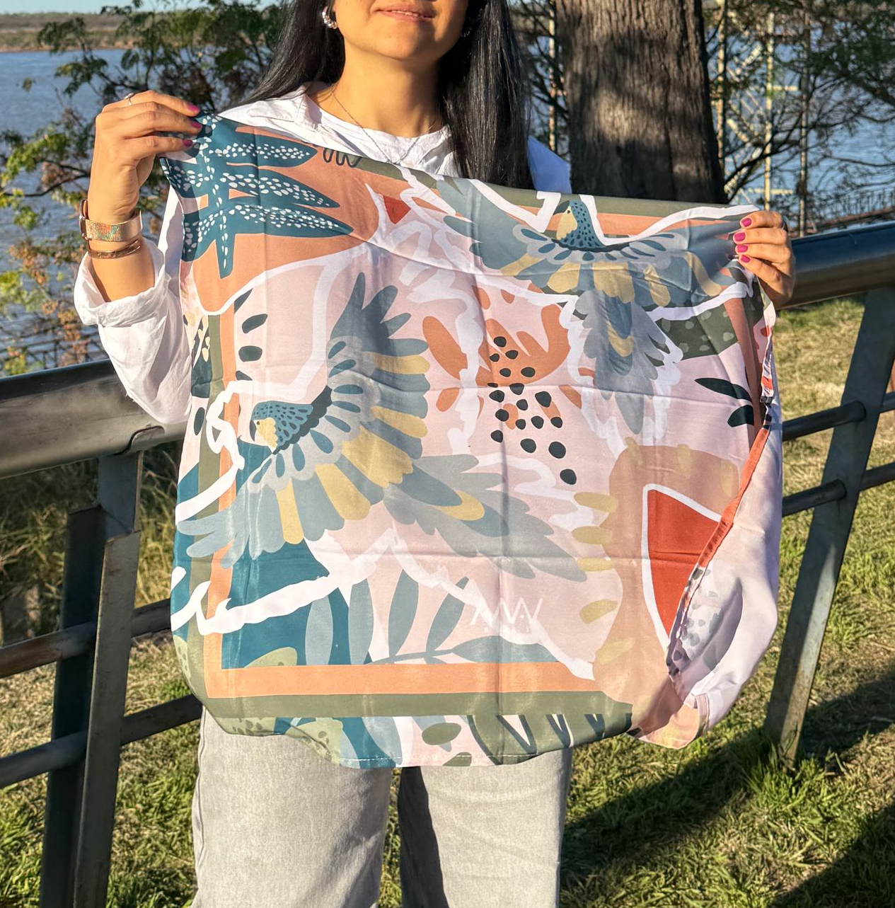 Mujer sosteniendo una bufanda con estampado floral al aire libre, con agua y árboles en el fondo.