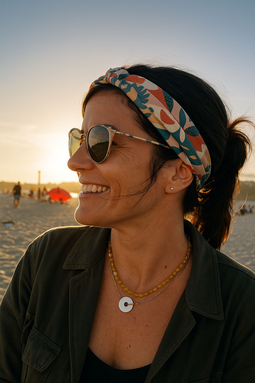 Mujer en una playa al atardecer, sonriendo, con gafas de sol, diadema, collar y una chaqueta verde.