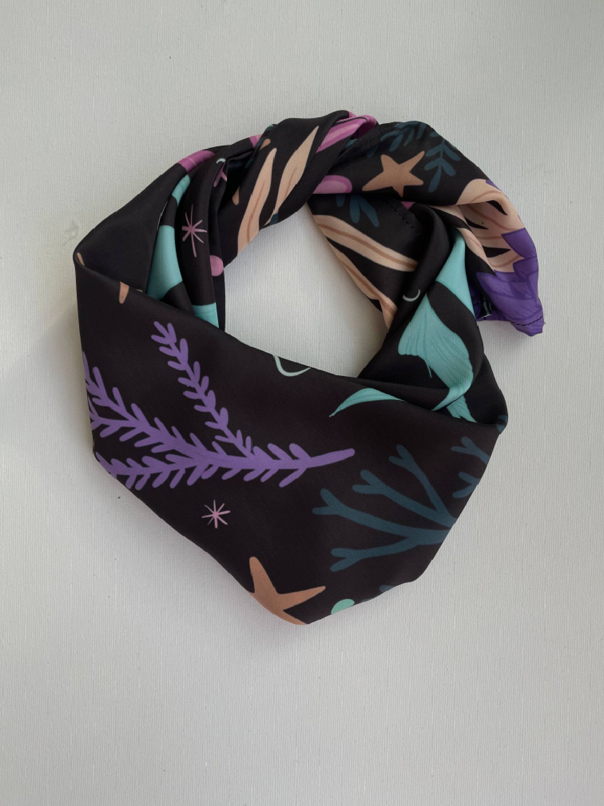 Bufanda infinita negra con estampado floral y de estrellas en verde azulado, morado y tostado sobre fondo blanco.