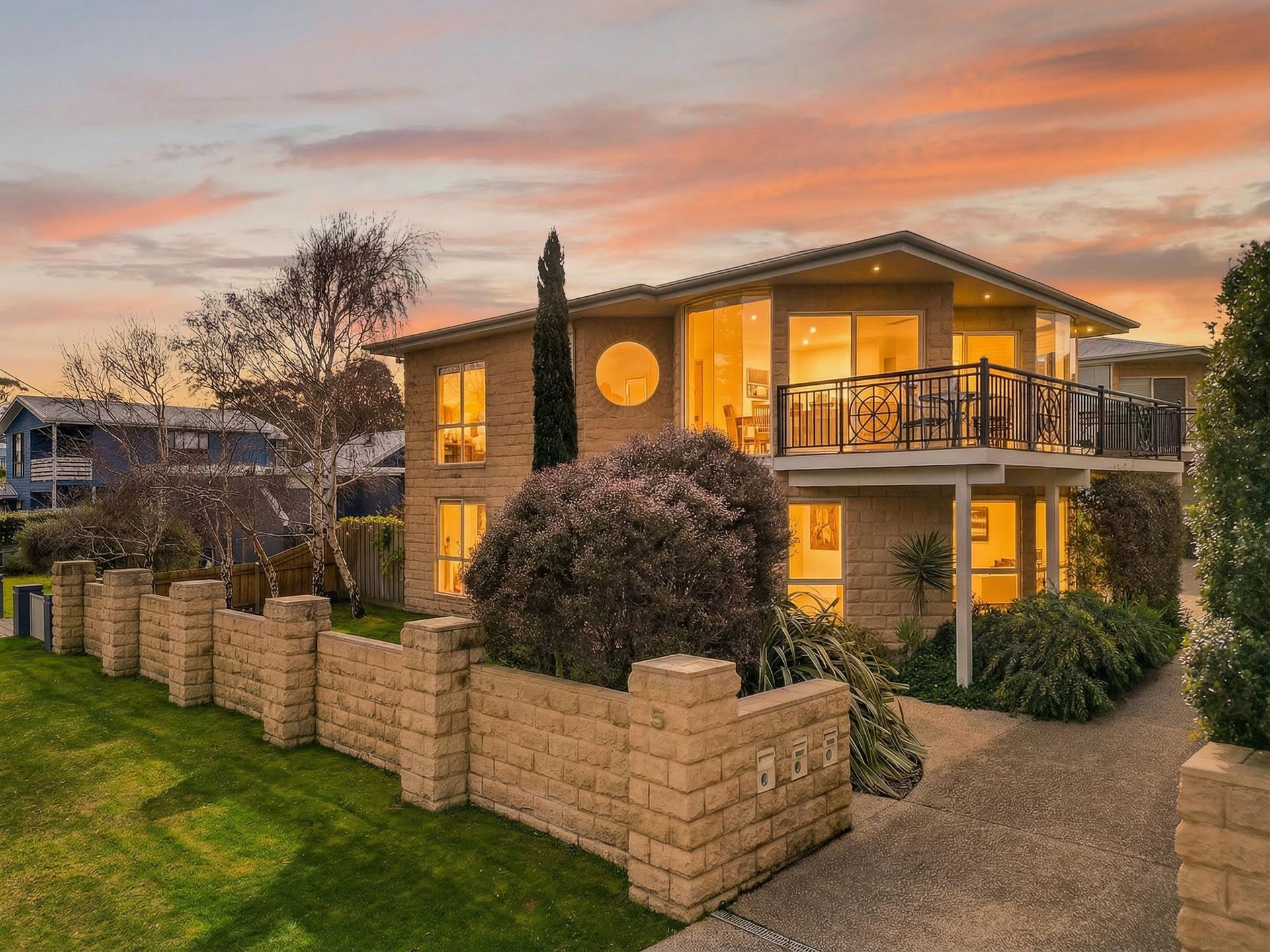 A two-story tan brick house with a balcony and a brick fence under a sunset sky.