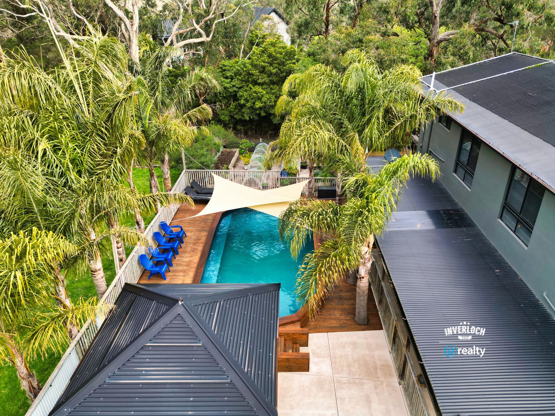 Aerial view of a swimming pool surrounded by a wooden deck and trees, next to a modern gray building.