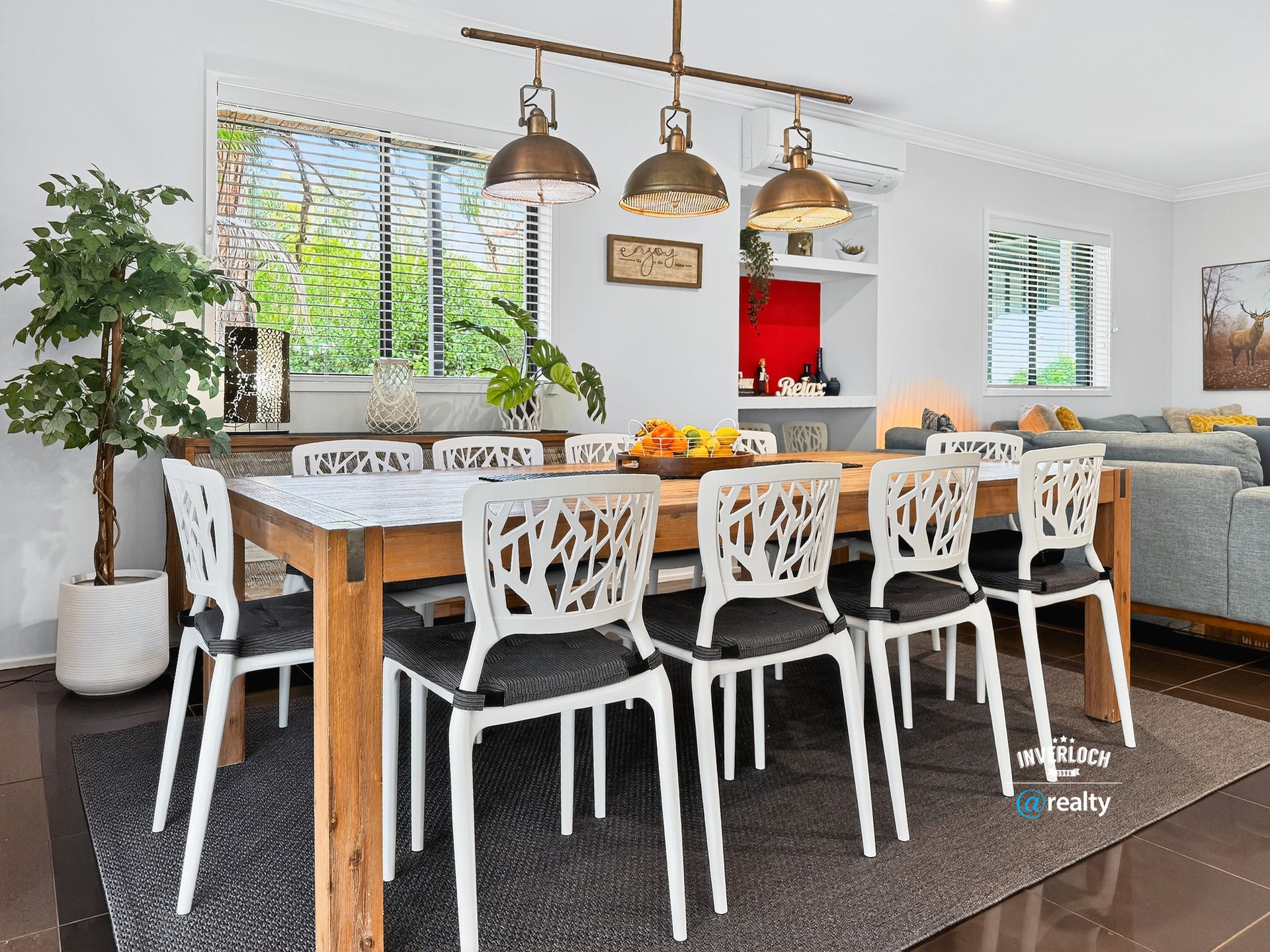 Dining room with wooden table, white chairs, and three pendant lights. Window with blinds, a plant, and built-in shelving.