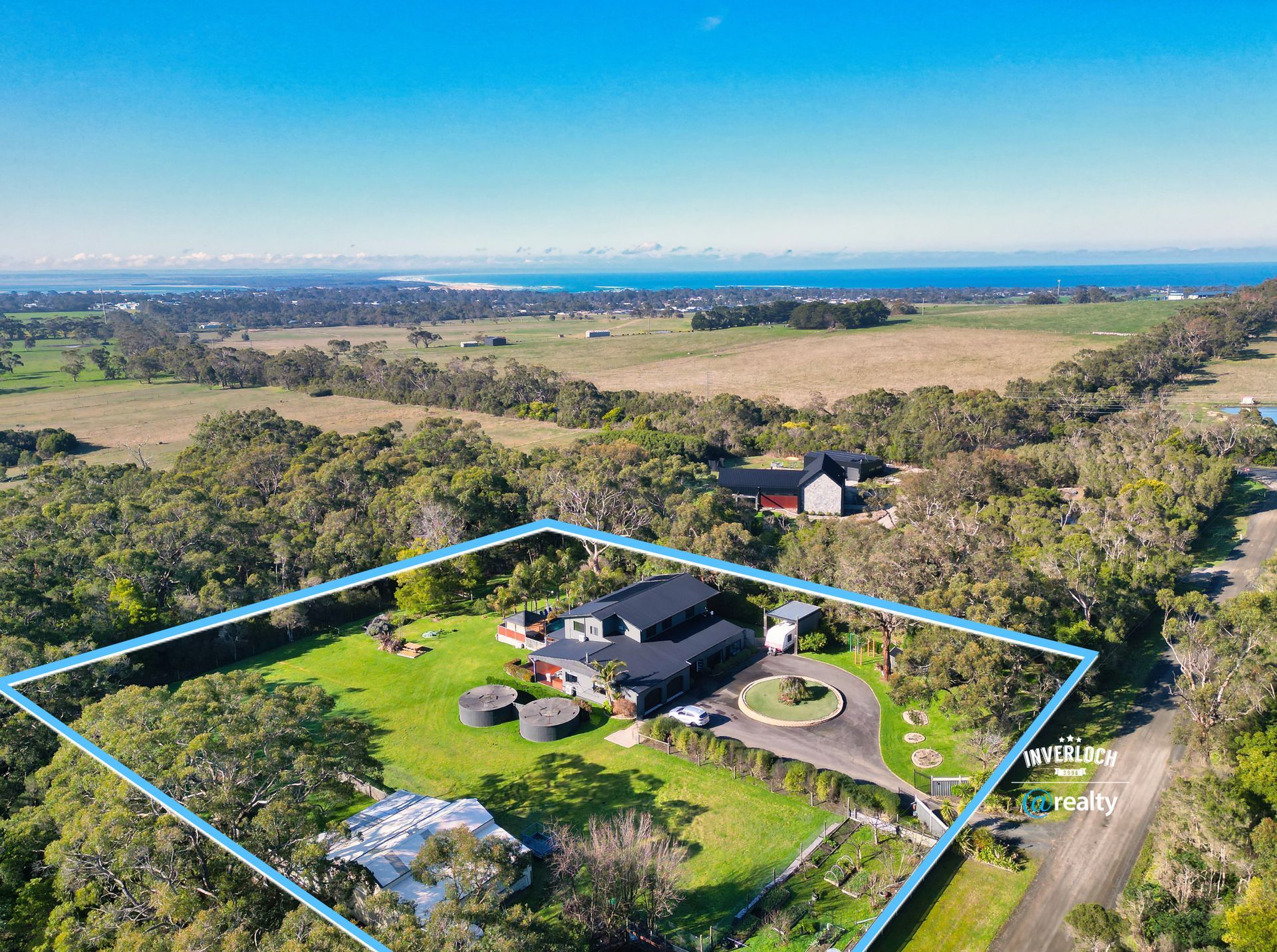 Aerial view of a large house with a circular driveway, surrounded by green fields and trees, near the ocean.