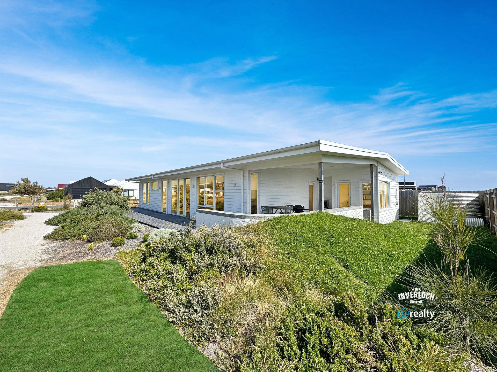 White coastal home with long veranda, green grass, and blue sky.