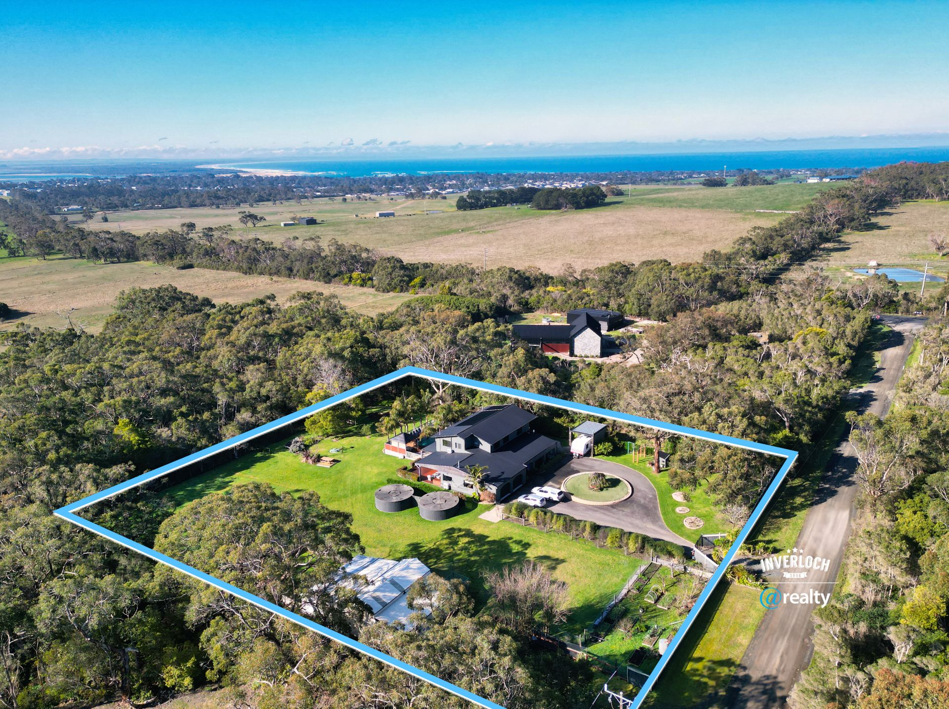 Aerial view of a black house with a circular driveway surrounded by trees and fields, with ocean in the background.