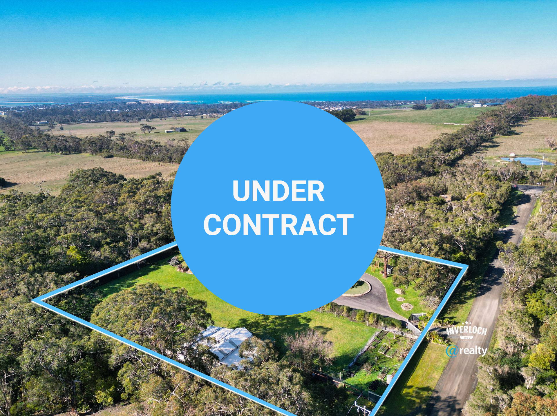 Aerial view of a modern black house surrounded by trees, fields, and the ocean in the background.