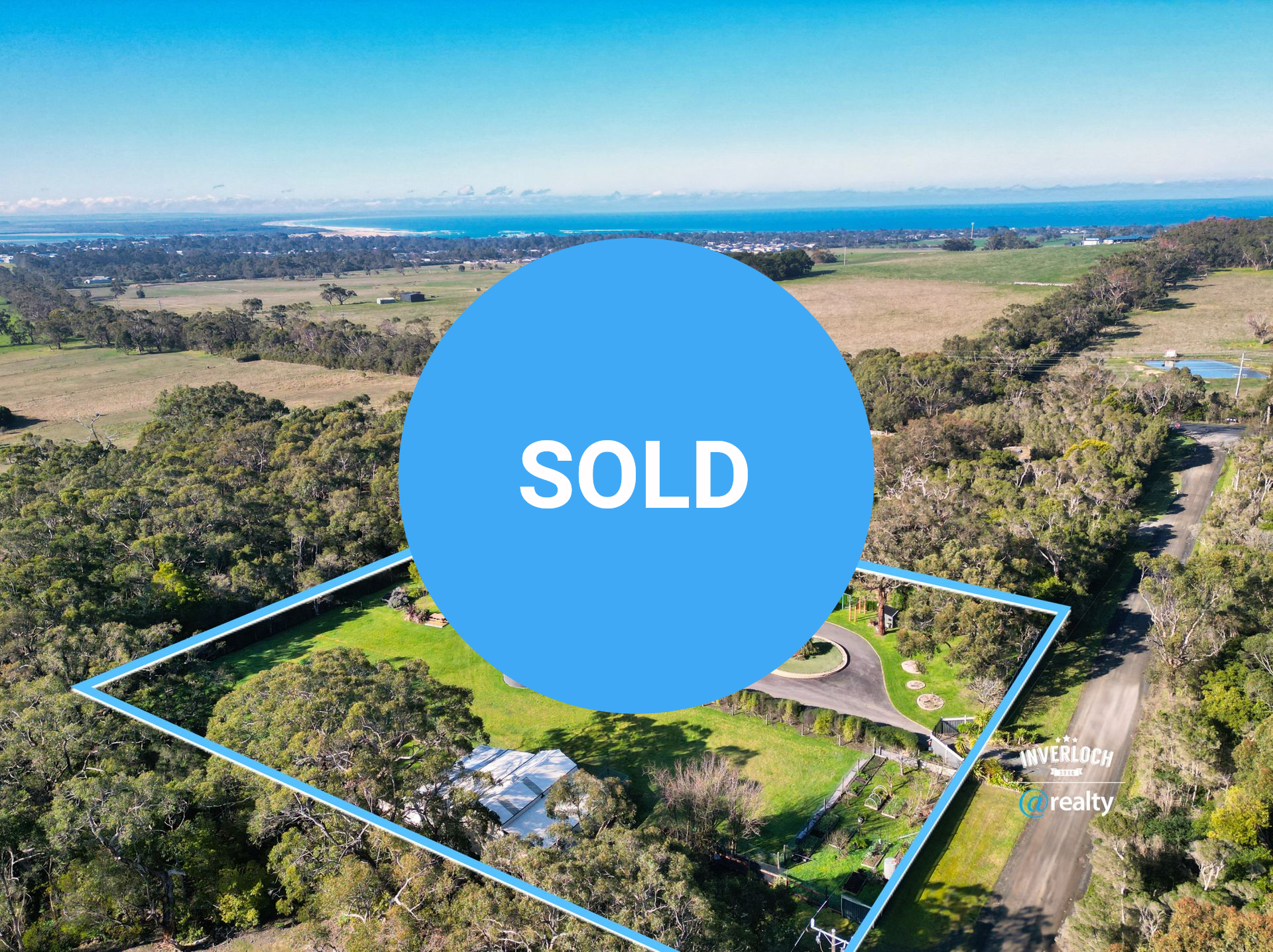 Aerial view of a black house with a circular driveway surrounded by trees and fields, with ocean in the background.