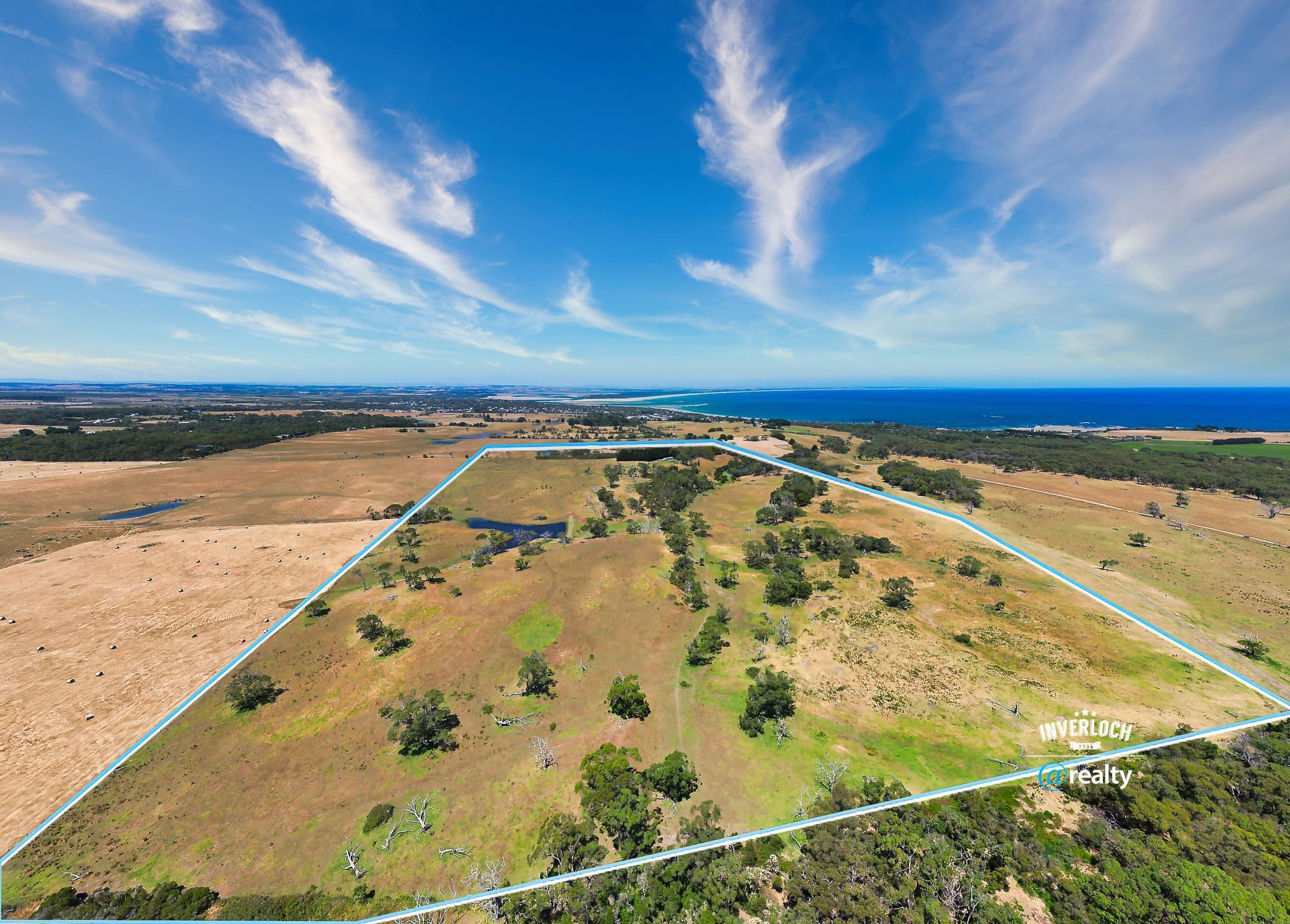 Aerial view of rural land with blue outline, trees, and ocean in the distance under a bright blue sky.