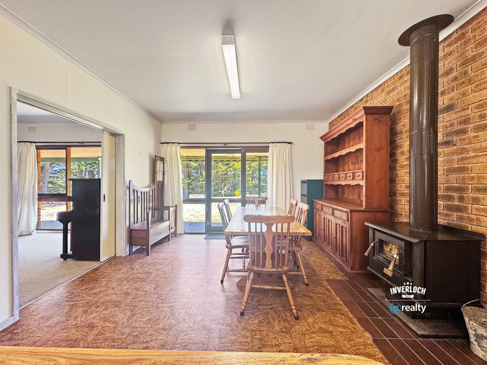Dining room with wooden table and chairs, brick fireplace, and hutch. Doorway leads to living room with piano.