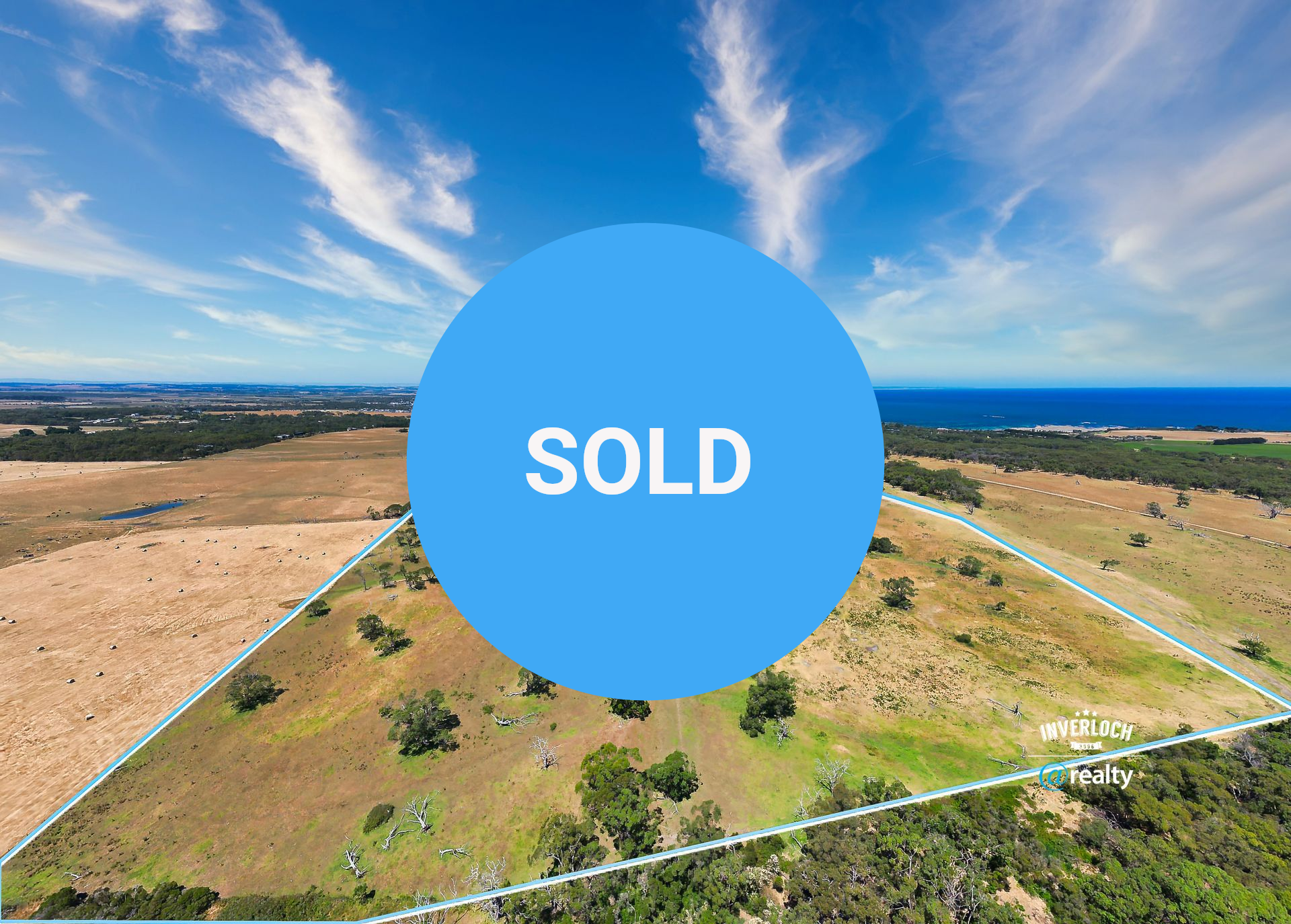 Aerial view of rural land with blue outline, trees, and ocean in the distance under a bright blue sky.