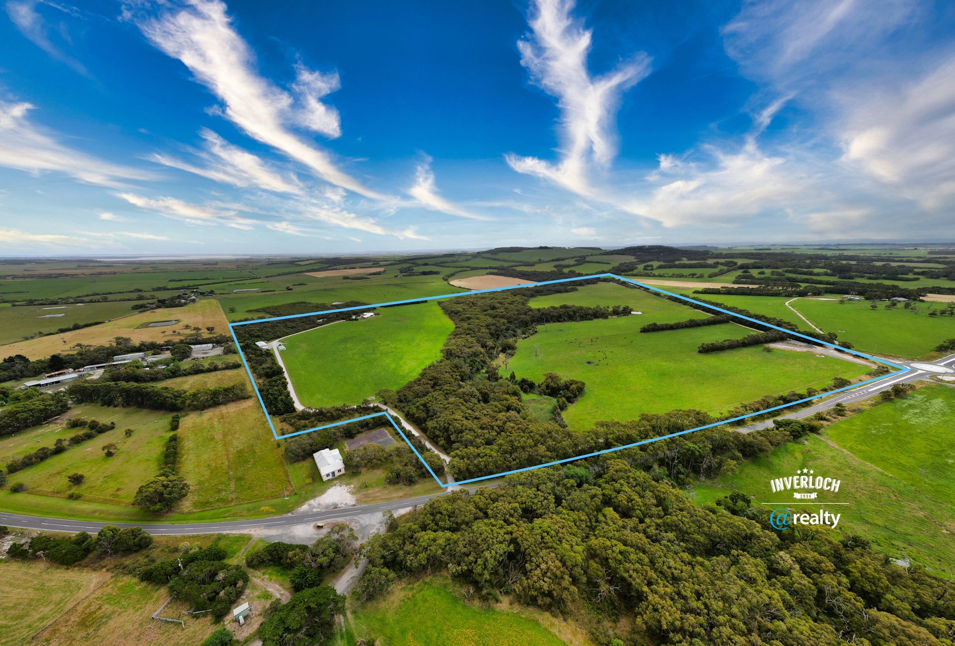Aerial view of a green field bordered by trees under a bright blue sky with white clouds.