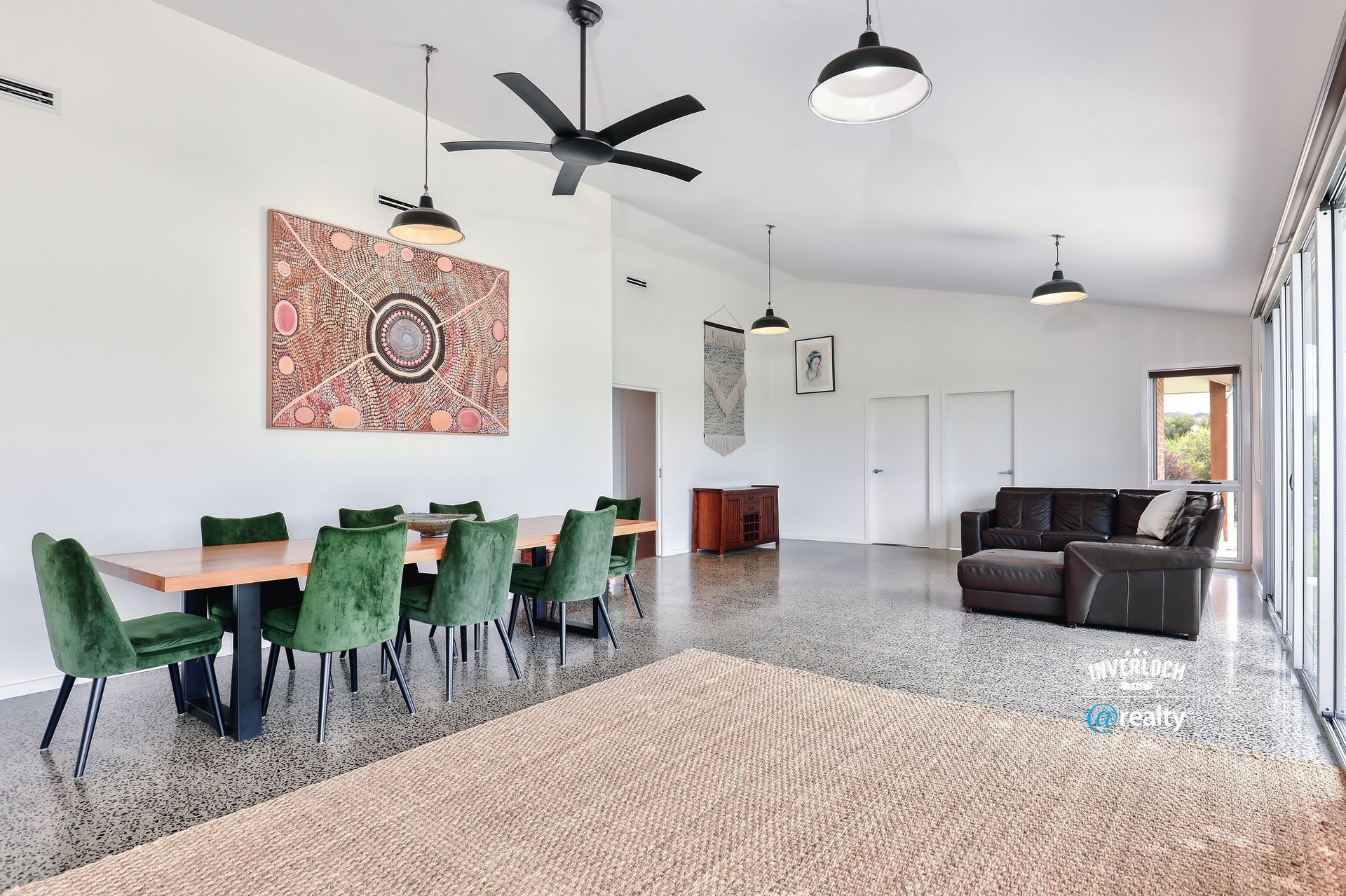 Spacious, modern living area with dining table, green chairs, and a brown sofa. Large rug on a speckled floor.