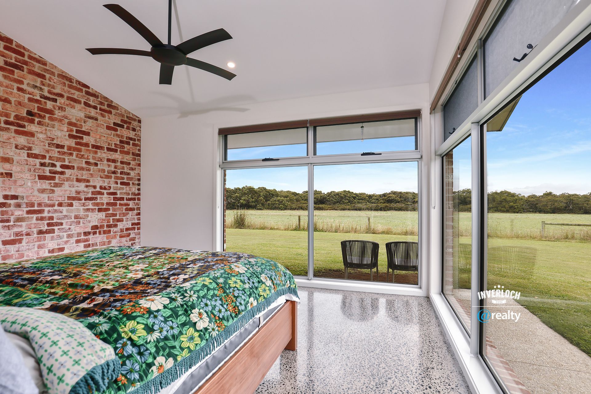 Bedroom with a brick wall, large windows overlooking a field, bed with a colorful quilt, and a ceiling fan.