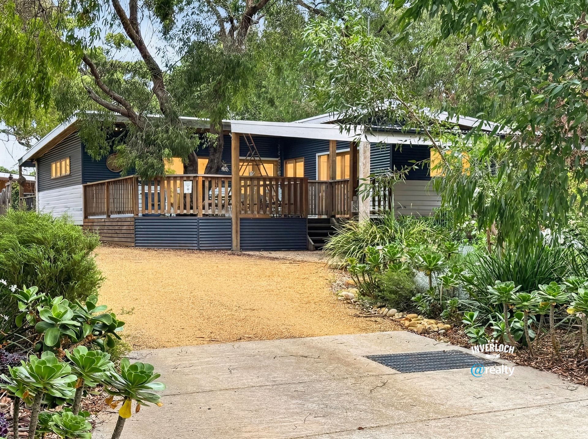 Wooden house with a porch and yellow gravel path, surrounded by trees and lush greenery.
