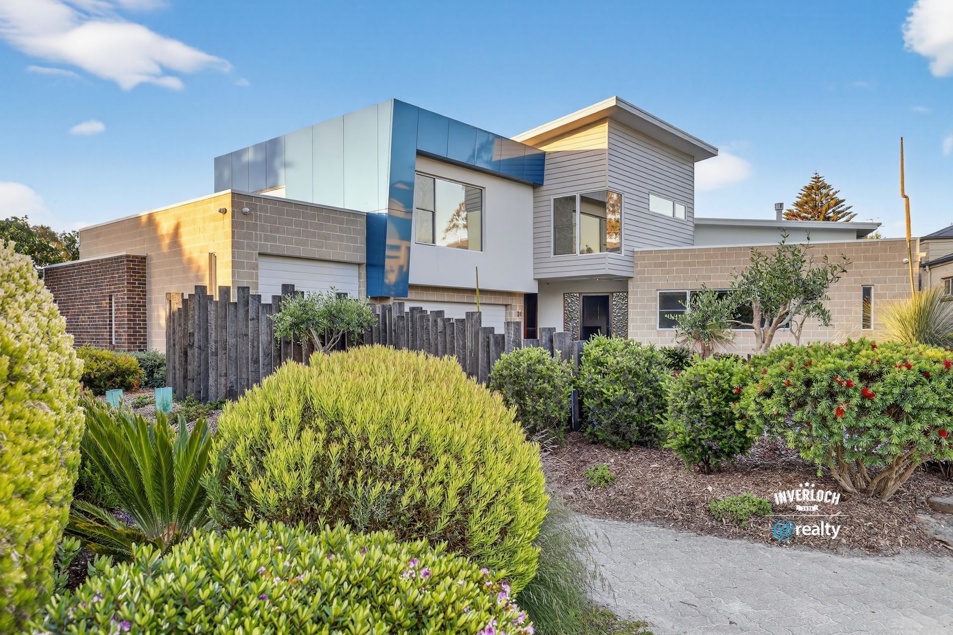 Modern house with blue and tan exterior, surrounded by green shrubs and a rustic wooden fence.