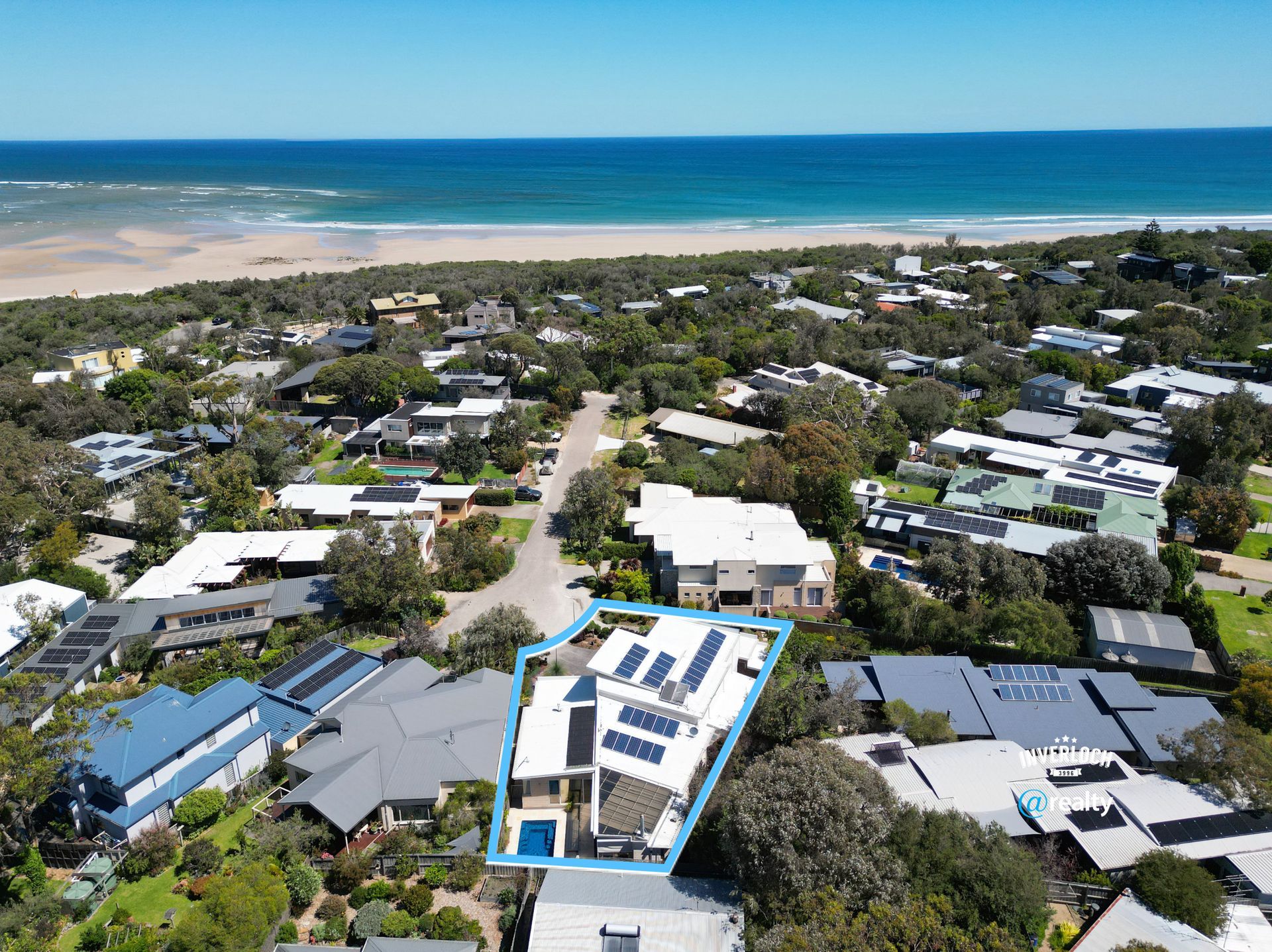 Coastal house on a dune, surrounded by green trees under a bright blue sky with clouds.