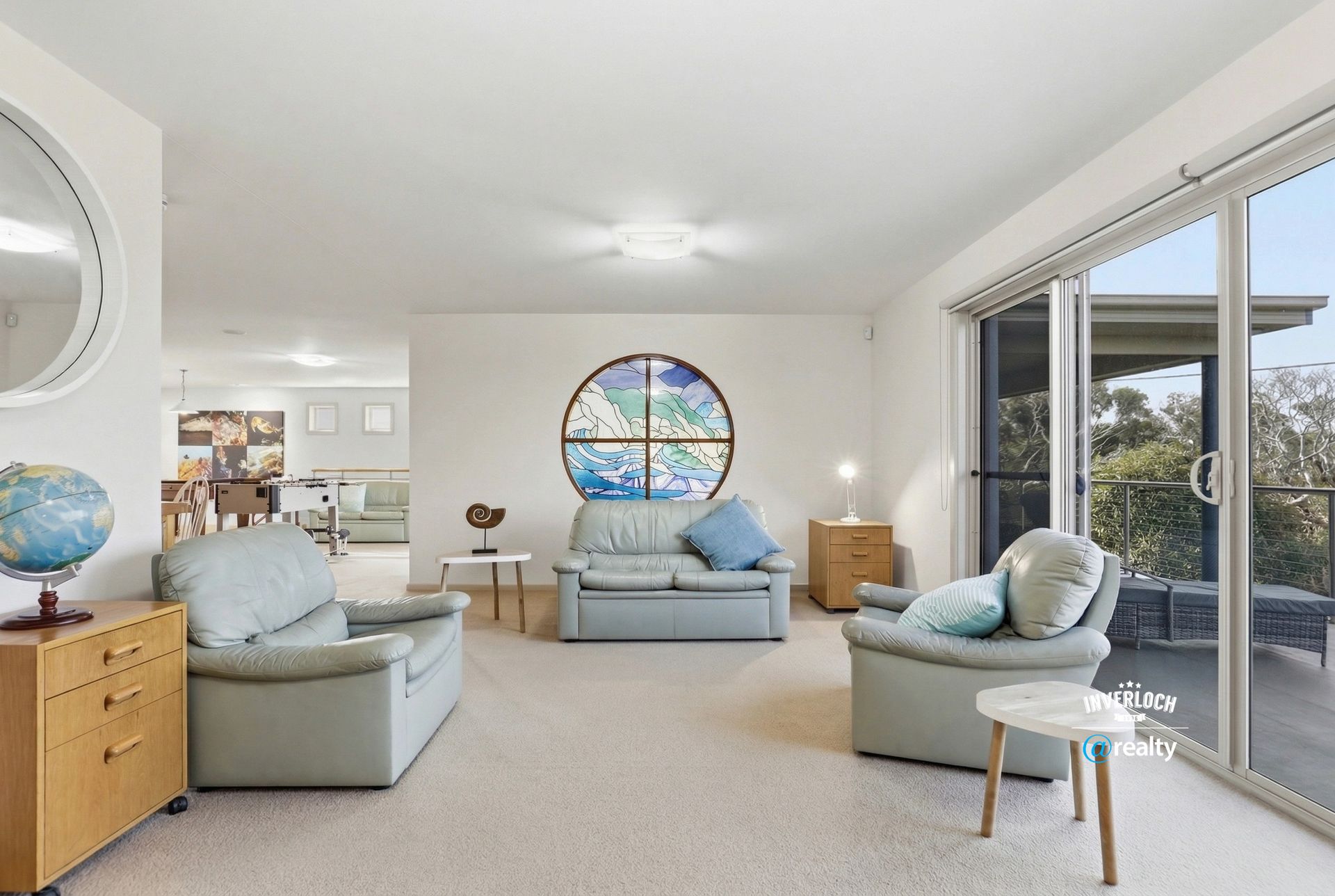 A bright living room with three light gray sofas, a globe on a wooden cabinet, and a large circular stained glass window.