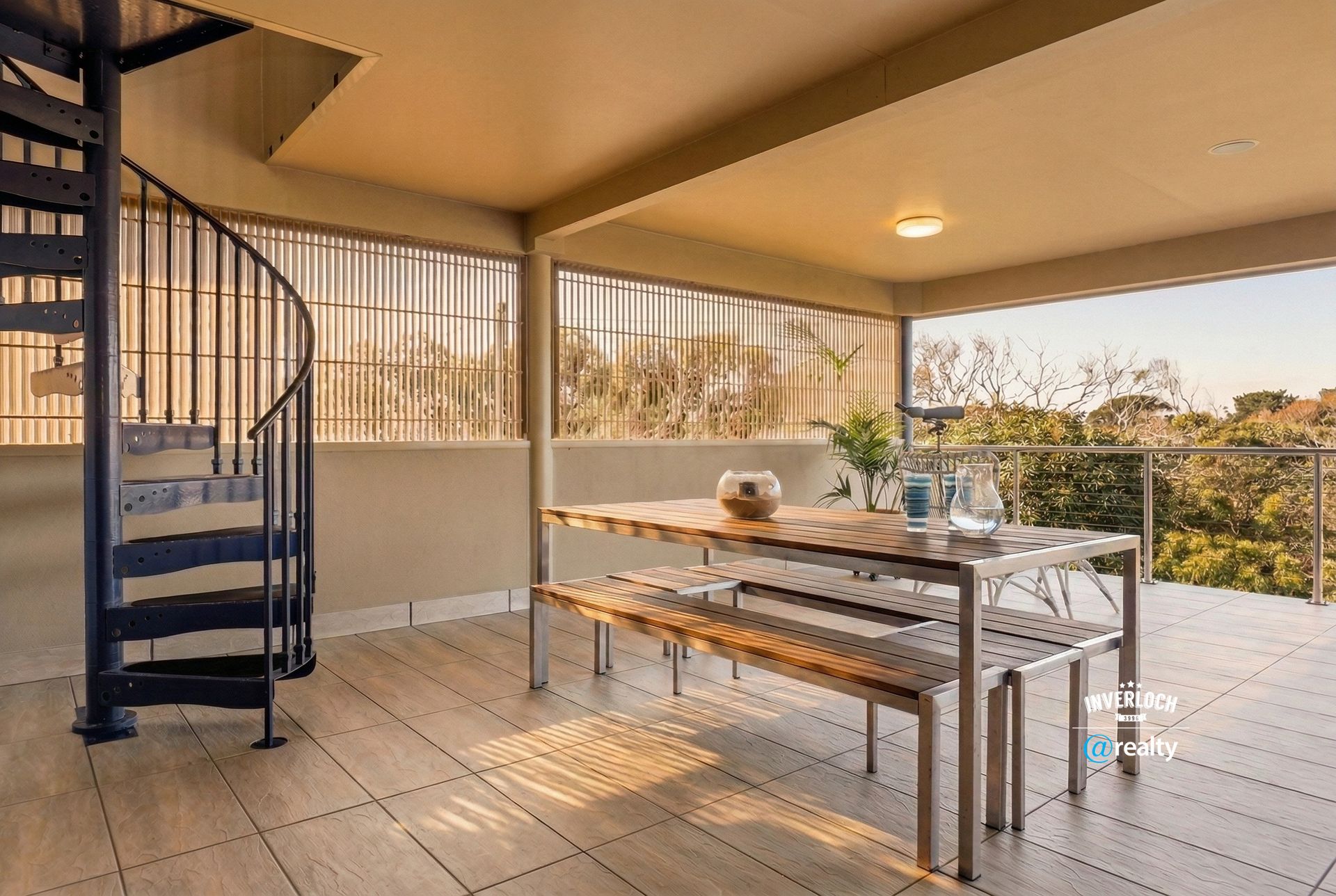 A covered outdoor patio featuring a wooden dining table, benches, and a dark spiral staircase overlooking trees.