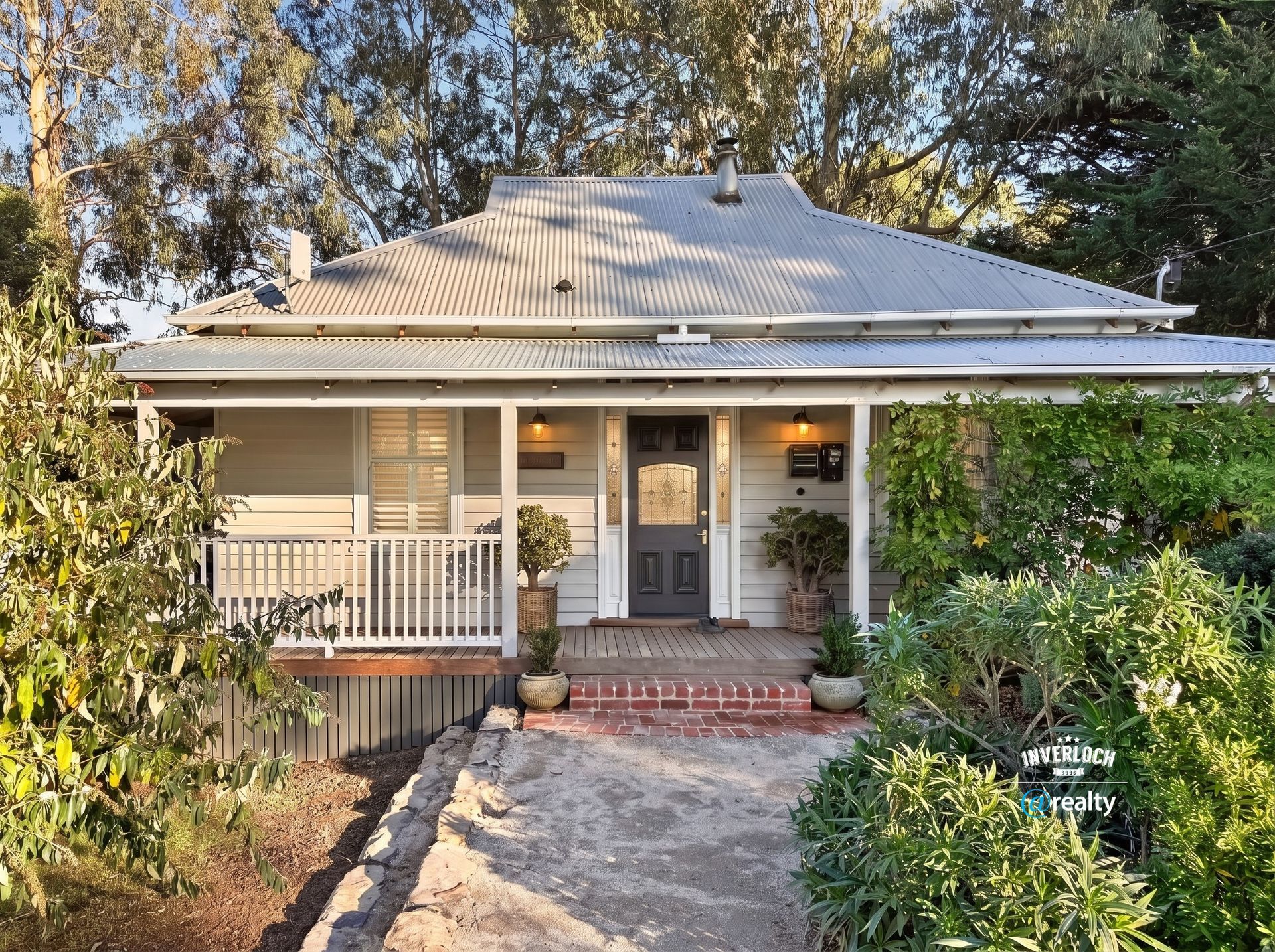 Front porch of a beige house with a white roof, brick steps, and lush greenery in the yard