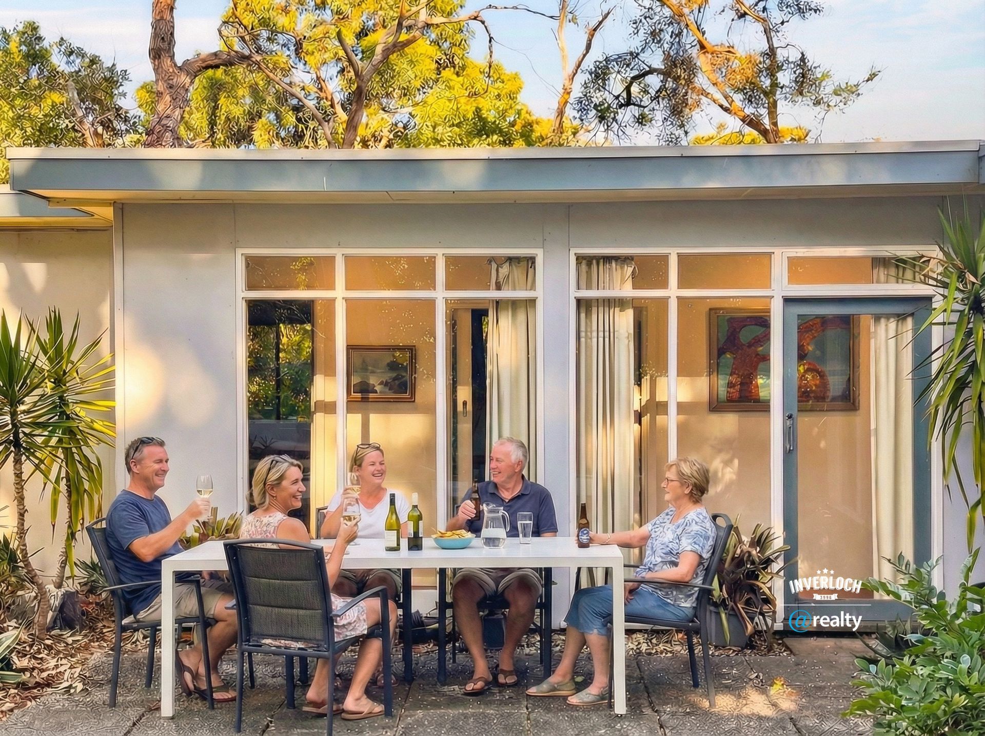 Five people sit around an outdoor table on a stone patio, smiling and drinking wine in front of a modern glass-walled home.