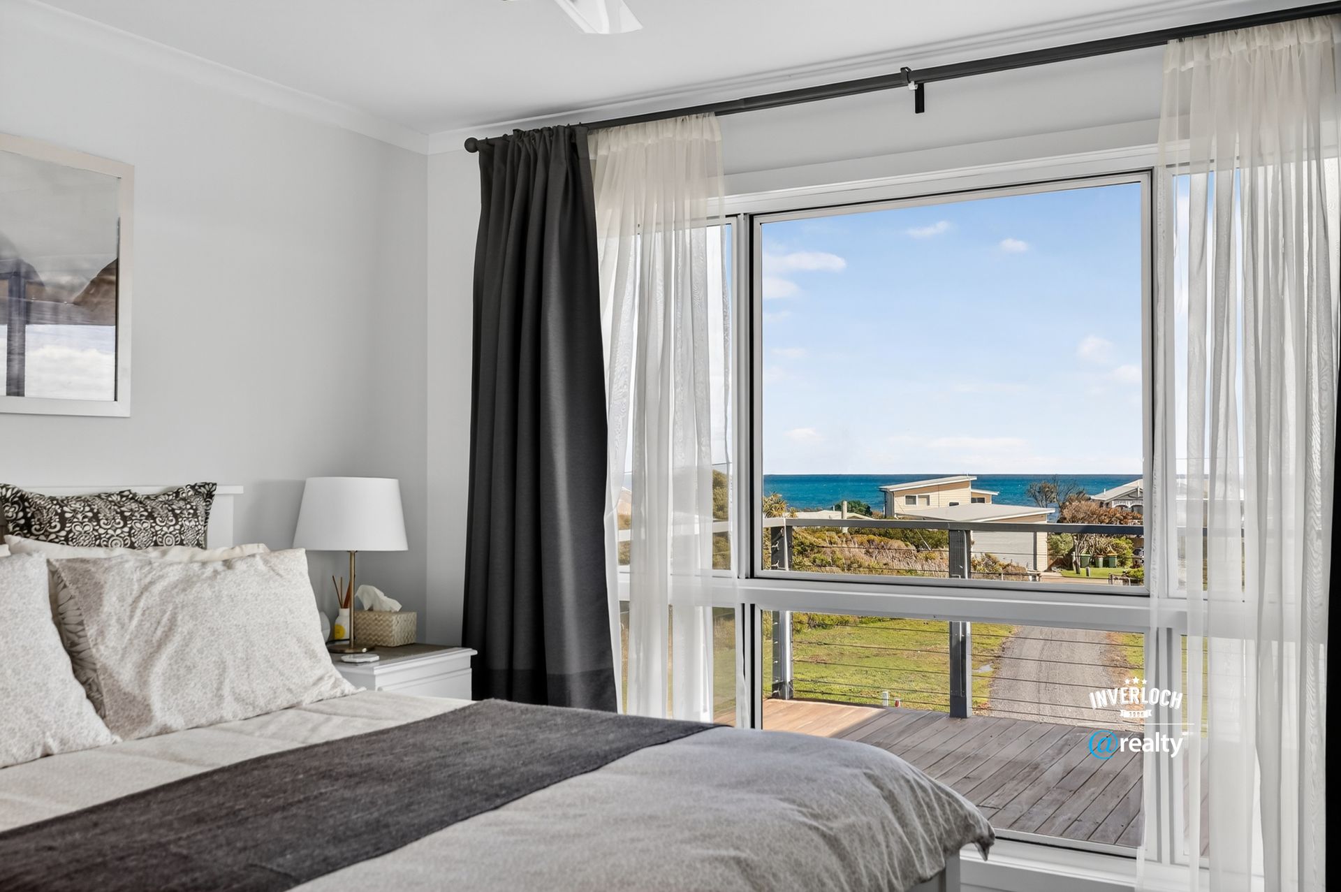 Bedroom with large window overlooking ocean; dark curtains, white bedding, seascape view.