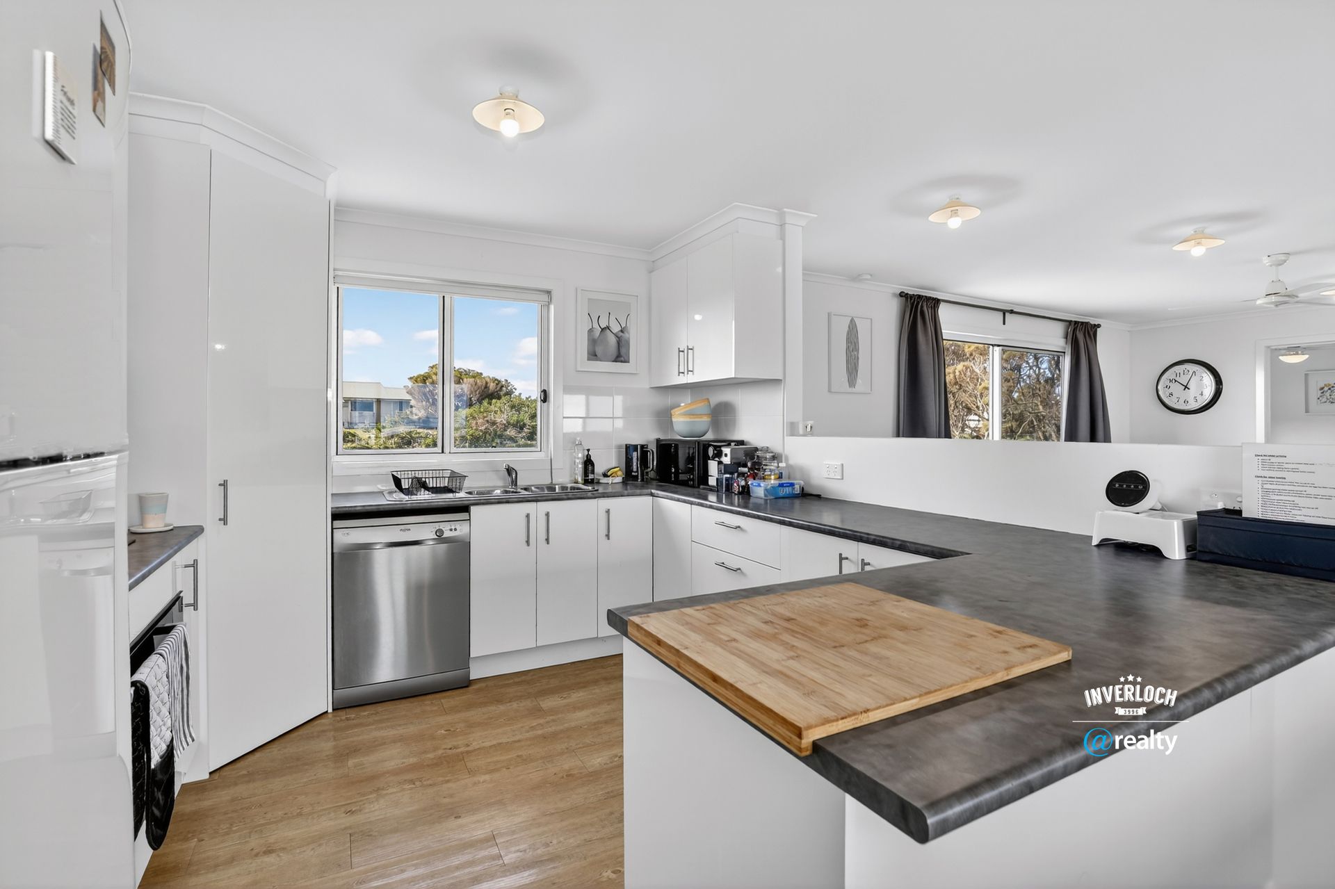 Bright white kitchen with stainless steel appliances, dark countertop, and a wooden cutting board.