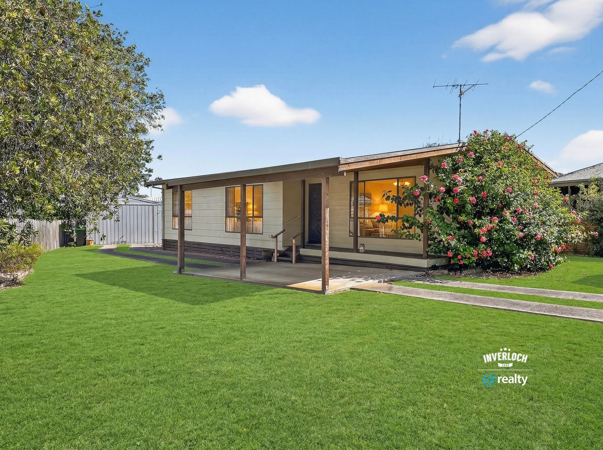 A single-story house with a covered patio, tan siding, and a large lawn under a blue sky with scattered clouds.